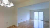 This single-floor space offers a clean, minimalist aesthetic defined by light hardwood floors that run throughout, reflecting natural light and lending warmth to the interior. The layout features an open-concept design, with a defined dining area transitioning seamlessly into a spacious living area. A classic pendant light fixture hangs in the dining zone, adding a subtle vintage touch to the otherwise neutral palette.

The living area is bathed in natural light, thanks to a full sliding glass door that leads to an outdoor balcony. This feature not only brightens the interior but also provides an easy indoor-outdoor connection, ideal for ventilation and enjoying outdoor views. The white walls throughout the space create a bright, neutral backdrop that maximizes the sense of openness.

Architectural details include a wide arched passageway between the dining and living areas, adding a touch of character while maintaining the flow of the open layout. White baseboards provide a crisp finish along the flooring. Overall, the space presents a light-filled, versatile environment well-suited for a variety of living arrangements.