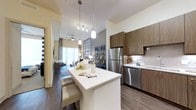 This single-floor residence showcases a thoughtfully designed open-concept layout that seamlessly connects the kitchen, dining, and living areas. The kitchen is a standout feature, equipped with a full suite of stainless steel appliances including a refrigerator, dishwasher, range, and microwave. Warm-toned, wood-grain cabinetry extends from floor to ceiling, providing generous storage, while crisp white countertops offer a clean contrast. A central island with matching cabinetry serves as both a prep space and a casual dining area, illuminated by stylish pendant lighting above. A textured tile backsplash and wood-look flooring complete the polished aesthetic.

The open living and dining area benefits from elevated ceilings, a ceiling fan, and large windows that allow natural light to fill the space. The layout accommodates both a dining table and a comfortable seating arrangement, creating a versatile and welcoming environment.

The bathroom features clean, contemporary finishes, including a wood-grain vanity with a white countertop, a large mirror framed by modern sconce lighting, and a tiled tub-shower combination. Neutral tile flooring and soft gray walls lend the space a calm, spa-like quality. Throughout the residence, consistent use of warm wood tones, neutral palettes, and modern fixtures creates a cohesive and refined atmosphere.