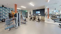 This modern fitness center is thoughtfully designed to accommodate a wide range of workout styles and fitness goals. The space is organized into distinct activity zones, each outfitted with professional-grade equipment. The cardio area features a row of treadmills, ellipticals, and a stair climber positioned along floor-to-ceiling windows that invite natural light and offer views of an outdoor pool area, creating an energizing atmosphere. Ceiling fans overhead add to the comfort of the space.

The strength and free weights section includes adjustable benches, a dumbbell rack, and Matrix selectorized machines, complemented by large wall mirrors that enhance the sense of openness. A dedicated cycling studio houses multiple stationary bikes, each equipped with individual screens, and is anchored by a wall-mounted television for group viewing.

A functional training zone is highlighted by a bold geometric-patterned accent wall in teal and blue tones, providing a striking visual backdrop. This area is equipped with a multi-tier rack stocked with medicine balls, stability balls, and kettlebells, as well as battle ropes and a BOSU ball for dynamic training. Built-in cubbies offer convenient storage for accessories.

Consistent neutral-toned flooring, bright linear lighting, and clean white walls throughout give the fitness center a polished, professional feel. A water fountain station and accessible entry doors complete this well-appointed facility.
