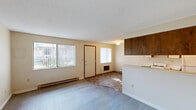 Located at 1116 E 30th Ave in Spokane, WA, this single-floor, one-bedroom unit offers a functional and straightforward layout. The combined kitchen and living room serves as the heart of the space, with the kitchen featuring a galley-style design equipped with white cabinetry, dark wood upper cabinet accents, a coil-burner electric stove, a refrigerator, and a single-basin sink with light-toned countertops. A peninsula separates the kitchen from the living area, which is finished with low-pile carpet and a textured ceiling. Multiple windows fitted with vertical blinds bring in natural light, and baseboard heating runs along the exterior walls throughout. The bedroom is a generously sized, carpeted room with a large window and its own baseboard heating unit. A hallway closet with sliding wood-paneled doors provides ample storage. The bathroom, accessible from the bedroom hallway, includes a toilet, vanity, and tile-style flooring. The overall aesthetic is clean and utilitarian, with warm wood tones offering contrast against neutral cream-colored walls, making this a practical and comfortable living space in a residential Spokane neighborhood.