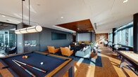 This upscale amenity lounge is thoughtfully designed across a single floor, blending contemporary style with warm, inviting materials. The space is defined by striking wood-slat ceilings accented with recessed and cove lighting, creating a refined ambiance throughout. Hardwood flooring transitions seamlessly into plush area rugs, delineating distinct zones within the open layout.

The main lounge area features multiple curated seating arrangements, including deep leather sofas, tufted settees, and accent chairs in rich cognac and earthy tones, all centered around low-profile coffee tables. Floor-to-ceiling windows wrap around the perimeter, flooding the interior with natural light and offering expansive panoramic city views.

A dedicated game area houses a full-sized billiards table beneath a sculptural pendant light, framed by the same sweeping windows showcasing the skyline. Nearby, a built-in wood shelving unit with integrated television serves as a focal point for a more intimate lounge nook.

An entry foyer sets an elegant tone with a bold abstract artwork, a sculptural globe chandelier, and a seating vignette of vibrant blue accent chairs. Large-format projection or display screens are integrated into the darker accent walls, making the space equally suited for social gatherings or presentations. The overall design balances functionality with a polished, contemporary aesthetic.