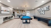 The Cue Gameroom at Savoye is a 540-square-foot amenity space designed to deliver an engaging and social recreational experience. The room's contemporary aesthetic is defined by soft gray walls, light wood-look flooring, and a curved accent wall adorned with decorative natural wood slice wall art, adding an organic, artistic touch to the space. A bold marquee-lit "GAME ON" sign serves as a focal statement on one wall, setting the tone for the room's energetic atmosphere. At the center of the room stands a striking dark-framed pool table with vibrant blue felt, illuminated by a modern branching chandelier overhead. A full-length shuffleboard table runs along one side, complementing the room's lineup of games. An arcade game cabinet provides additional entertainment options. Recessed lighting and the chandelier ensure the space is well-lit throughout. High-top tables with sleek black bar stools offer casual seating for spectators and socializers. A built-in counter with bar stools along one wall provides a workspace or bar-style seating area, accompanied by a dedicated cue rack. Large windows along the curved wall allow natural light to filter in, enhancing the room's open and inviting ambiance.
