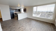 Located at 4351 15th Ave S in Seattle's 98108 zip code, this single-floor unit offers a thoughtfully designed open-concept living space. The kitchen is outfitted with dark espresso cabinetry, granite-style countertops, and stainless steel appliances — including a refrigerator, range, and microwave — all arranged around a peninsula that seamlessly connects the kitchen to the living area. Light-toned wood-look flooring runs throughout, lending warmth and continuity across the space. Large windows with horizontal blinds allow generous natural light to fill the interior, while also framing views of the surrounding Seattle neighborhood and skyline beyond. The clean white walls and modern flush-mount lighting fixtures contribute to a bright, minimalist aesthetic. The open layout between the kitchen and living area creates a spacious, airy feel well-suited for both everyday living and entertaining. The overall design balances contemporary style with practical functionality, making it an efficient and attractive urban living space in South Seattle.