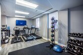 This fitness center in Memphis, Tennessee offers a thoughtfully designed workout environment spread across a single floor. The space combines functional fitness infrastructure with polished interior design, making it an inviting destination for exercise.

Upon entering through a glass-paneled door, the main area is immediately distinguished by a striking amber bubble chandelier that adds an unexpected decorative elegance to the workout environment. Light wood-look flooring runs throughout, complementing the soft gray walls and lending the space a clean, modern aesthetic.

The cardio zone is well-appointed with multiple True-brand treadmills, elliptical trainers, and a recumbent bike, all positioned beneath recessed panel ceiling lighting and a wall-mounted flat-screen television for entertainment during workouts. Natural light filters in through windows along the perimeter, enhancing the open and airy feel of the space.

A dedicated strength and free weights area is tucked toward the rear, featuring a multi-function cable machine with an adjustable bench, a rack of medicine balls, dumbbells, and stability balls, all set on protective rubber matting. A standing scale near the entrance provides a convenient wellness tracking station. The facility also includes safety essentials such as a fire extinguisher, and benefits from direct exterior access, connecting seamlessly to the surrounding property.