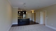 This single-floor residence offers a thoughtfully designed open-concept layout that blends style and functionality. The kitchen serves as a central focal point, featuring rich dark cabinetry, granite countertops, and a curved island that provides both workspace and a natural gathering spot. Stainless steel appliances, including a refrigerator, microwave, and range, add a contemporary edge, while a trio of pendant lights suspended above the island creates warm, focused illumination. Crown molding runs throughout the space, lending an air of refined detail to the ceilings. The kitchen transitions seamlessly into the living area, which is finished with plush carpeting that adds comfort underfoot, contrasting elegantly with the dark hardwood-style flooring in the kitchen. Recessed lighting and wall-mounted sconces complement the natural warmth of the color palette, which features soft, neutral tones on the walls. Several interior doors lead to additional rooms, suggesting a well-organized floor plan with dedicated private spaces. The overall ambiance is modern yet welcoming, with a careful balance of dark and light elements that give the space a polished, cohesive character throughout.