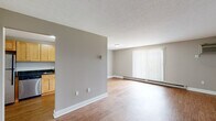 This single-floor unit is designed with a practical, open layout that maximizes the available living space. Throughout the unit, warm-toned hardwood-style flooring ties the rooms together, complemented by neutral grey walls and crisp white baseboards and trim that lend a clean, cohesive aesthetic.

The living area is generously proportioned, featuring a large window fitted with vertical blinds that allows natural light to fill the space. A baseboard heating system runs along the perimeter, ensuring year-round comfort. The semi-open layout connects the living area to the kitchen via a partial dividing wall.

The kitchen is well-equipped, featuring light maple cabinetry with ample upper and lower storage, dark granite-style countertops, and a full suite of stainless steel appliances including a refrigerator, dishwasher, and electric range.

The bedrooms are comfortably sized, each with hardwood-style flooring, neutral walls, and baseboard heating. One bedroom includes a walk-in closet with built-in shelving, while another features a window with outdoor views and a wall-mounted air conditioning unit.

The unit includes two bathrooms: a full bathroom with a white vanity, mirror, and baseboard heater, and a separate water closet. The overall ambiance is bright, clean, and move-in ready.