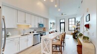 Located in the heart of historic Charleston, South Carolina, this thoughtfully designed one-bedroom, three-bathroom residence offers a refined blend of modern finishes and timeless architectural character. Soaring ceilings throughout amplify the sense of space, while large black-framed grid windows flood the interior with abundant natural light and frame picturesque views of the Charleston skyline and surrounding greenery.

The open-concept kitchen and living area form the heart of the home. The kitchen is outfitted with white shaker cabinetry, a textured geometric tile backsplash, stainless steel appliances, and light quartz countertops. A white center island with an X-base provides casual seating and additional workspace. The adjacent living room features a ceiling fan, direct balcony access through a striking black-framed glass door, and light hardwood flooring that flows throughout the unit.

The bedroom is a serene retreat with soaring walls, a walk-in closet with built-in shelving, and a large window that invites natural light. Three well-appointed bathrooms feature white cabinetry, tile flooring, and clean, minimalist finishes. A hallway connects the living spaces efficiently, completing this cohesive and stylish Charleston residence.