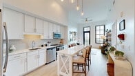 Located in the heart of historic Charleston, South Carolina, this thoughtfully designed one-bedroom, three-bathroom residence offers a refined blend of modern finishes and timeless architectural character. Soaring ceilings throughout amplify the sense of space, while large black-framed grid windows flood the interior with abundant natural light and frame picturesque views of the Charleston skyline and surrounding greenery.

The open-concept kitchen and living area form the heart of the home. The kitchen is outfitted with white shaker cabinetry, a textured geometric tile backsplash, stainless steel appliances, and light quartz countertops. A white center island with an X-base provides casual seating and additional workspace. The adjacent living room features a ceiling fan, direct balcony access through a striking black-framed glass door, and light hardwood flooring that flows throughout the unit.

The bedroom is a serene retreat with soaring walls, a walk-in closet with built-in shelving, and a large window that invites natural light. Three well-appointed bathrooms feature white cabinetry, tile flooring, and clean, minimalist finishes. A hallway connects the living spaces efficiently, completing this cohesive and stylish Charleston residence.