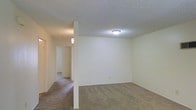 This single-floor space offers a straightforward and functional layout designed for comfortable everyday living. The main living area is finished with wall-to-wall carpeting in a warm neutral tone, providing a soft and inviting underfoot feel throughout. Smooth, off-white walls create a clean, bright backdrop that allows for versatile interior styling. A textured popcorn ceiling adds a classic residential character to the space, with a flush-mounted ceiling light fixture providing ambient illumination.

A structural column serves as a subtle room divider, defining the transition between the main living area and the adjoining hallway. The hallway extends toward additional rooms, hinting at a well-organized floor plan with clear separation between living and private spaces. Wall-mounted sconce lighting in the hallway adds a warm, welcoming glow to the corridor.

The neutral color palette and consistent flooring throughout the space contribute to a cohesive and unified aesthetic. The open feel of the main room, combined with the connected hallway layout, makes the space feel practical and easy to navigate. Overall, this single-level space presents a clean, move-in-ready environment well-suited for a variety of living arrangements.