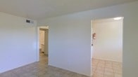 This single-story space presents a straightforward, functional layout characterized by its clean aesthetic and neutral palette. The interior is finished with light beige ceramic tile flooring that extends consistently throughout the visible areas, contributing to a cohesive and easy-to-maintain environment. Walls are painted in crisp white, enhancing the sense of brightness and openness across the space.

The main area is defined by a central dividing wall that creates a natural separation between zones, with open doorways on either side providing access to adjoining rooms. To the left, a doorway leads into what appears to be a bathroom, where a toilet and additional tile flooring are visible. To the right, a hallway extends further into the unit, lined with white walls and equipped with a mounted fire extinguisher, indicating safety compliance.

The ceilings feature a subtle textured finish and are fitted with a visible air vent, suggesting a functional HVAC system. The overall ambiance of the space is clean, minimal, and move-in ready, with a no-frills design that prioritizes practicality. The uniform color scheme and consistent flooring materials give the interior a tidy, well-maintained character throughout.
