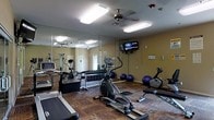 This fitness center is designed to support a well-rounded exercise routine within a single-floor layout. The room features a functional selection of cardio equipment, including treadmills, elliptical trainers, and a stationary bike, making it suitable for a variety of aerobic workouts. A dedicated free weight area includes a rack of dumbbells in graduated sizes, offering options for strength training. Exercise balls in multiple sizes are positioned along one wall, adding versatility for core and flexibility work. The flooring features a durable wood-patterned surface suited for high-traffic fitness use. Mirrored walls span a significant portion of the room, visually expanding the space and allowing users to monitor their form during exercise. Overhead ceiling fans provide continuous air circulation, contributing to a more comfortable environment during workouts. Fluorescent lighting ensures the room is well-lit throughout. A wall-mounted flat-screen television is positioned for easy viewing from multiple equipment stations. The walls are painted in a warm neutral tone, and the overall layout is organized to allow clear pathways between equipment, promoting safe and efficient use of the space.