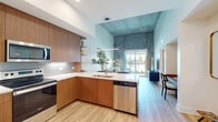 This thoughtfully designed single-floor residence showcases a seamless blend of contemporary architecture and warm, natural materials. The open-concept living area immediately impresses with its dramatically tall ceilings, where a striking textured concrete accent wall creates a bold focal point that extends upward, amplifying the sense of volume throughout the space. Light wood-tone flooring flows continuously from the kitchen through the living area, unifying the layout.

The kitchen is well-appointed with rich wood cabinetry extending to the upper reaches of the walls, complemented by clean white countertops, stainless steel appliances, and an L-shaped peninsula with bar seating. Open shelving adds a modern, functional touch. The adjacent living area benefits from an abundance of natural light and large sliding glass doors that open to a private balcony overlooking a resort-style pool flanked by palm trees.

The bedroom continues the textured concrete accent wall detail, creating a cohesive design language throughout the residence, while a hallway connects to a dedicated laundry area and storage. The bathroom features light wood-look flooring, a glass-enclosed shower, and clean, minimal finishes. A separate area accommodates a home office or dining space, enhancing the unit's versatility.
