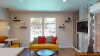 Located at 2376 Fairfield Ave in Fairfield, California, this single-floor community clubhouse is a well-appointed gathering space designed with a modern, eclectic aesthetic. The interior is unified by warm hardwood-style flooring, recessed lighting, and a distinctive teal-painted ceiling that adds a pop of color and visual interest throughout.

The lounge area serves as the social centerpiece, featuring large windows that frame views of an outdoor pool and seating area, flooding the space with natural light. A bold, textured accent wall with a distressed, multicolor mural adds an artistic focal point, complemented by a wall-mounted flat-screen television and floating wood shelves for decorative display.

Adjacent to the lounge is a functional kitchenette area highlighted by a white quartz-topped island with industrial-style bar stools, ideal for casual dining or socializing. A striking navy blue hexagonal tile backsplash frames the countertop wet bar area, while a mid-century modern brass chandelier with globe pendants provides warm, ambient lighting overhead. Decorative round mirrors and open shelving complete the contemporary look.

The space also includes a foosball table, offering a fun recreational element for residents. A direct exterior door provides easy access to the outdoor areas. Overall, this clubhouse strikes a balance between stylish design and practical amenity, making it an inviting hub for community gatherings.