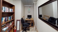 This single-floor space is thoughtfully arranged to support productivity and leisure reading in a quiet, functional environment. The library area is a standout feature, lined with large wooden bookshelves — including glass-doored cabinets — that house an extensive and well-organized collection of books spanning fiction, reference works, and more. A comfortable office chair with a cushioned seat is positioned nearby, creating a cozy reading nook within the shelving. The space also includes a dedicated computer workstation tucked into a compact alcove, furnished with a wooden desk, an ergonomic office chair, and a desktop monitor setup. A wall clock keeps the area task-oriented, while a security camera mounted near the ceiling reflects a practical attention to safety. The entry features a windowed door with a security light overhead, and the tile flooring throughout lends the space a clean, low-maintenance feel. Textured ceiling tiles and neutral-toned walls contribute to a calm, utilitarian atmosphere throughout. Whether used for research, quiet study, or casual browsing through a curated book collection, this space offers a well-equipped and organized environment for intellectual pursuits.