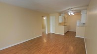 This single-floor space features an open-concept layout that seamlessly connects the main living area to the kitchen. The living room is finished with warm-toned hardwood flooring that extends throughout, creating a cohesive and inviting atmosphere. Neutral, light yellow walls reflect natural and artificial light effectively, contributing to a bright and airy feel. A ceiling fan with a light fixture is centrally mounted, providing both ventilation and overhead lighting.

The kitchen is positioned at the far end of the open living space and is equipped with white cabinetry, upper and lower units, and a sink integrated into the countertop. The white cabinetry contrasts cleanly against the warm wall tones. A passageway visible from the main area leads to an additional room, also equipped with a ceiling fan, suggesting further living or sleeping space beyond. A closed white door nearby likely provides access to a closet or utility area. White baseboards frame the perimeter of the rooms, adding a clean, finished look throughout the space.