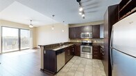 Located at 301 W Jefferson Blvd in downtown Fort Wayne, Indiana, this one-bedroom, one-bathroom residence offers a well-designed layout across a single living floor. The open-concept kitchen and living area serves as the heart of the unit, featuring rich dark cabinetry, granite countertops, a mosaic tile backsplash, and a full suite of stainless steel appliances including a range, built-in microwave, dishwasher, and refrigerator. A peninsula with bar seating and pendant lighting above creates a natural divide between the kitchen and living area, which is finished with wood-look flooring and a ceiling fan. Track lighting throughout the kitchen ensures ample illumination. The bedroom is a comfortable, carpeted retreat with a tray ceiling detail, large windows, and a sliding glass door that opens onto a private balcony overlooking a neighboring stadium — an ideal vantage point for sports enthusiasts. A dedicated closet provides convenient storage. The full bathroom includes a soaking tub and shower combination, a dark-vanity with granite countertop, and clean tile flooring that ties the space together cohesively. Natural light flows generously throughout the unit, enhancing its modern, urban aesthetic.