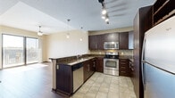 Located at 301 W Jefferson Blvd in downtown Fort Wayne, Indiana, this one-bedroom, one-bathroom residence offers a well-designed layout across a single living floor. The open-concept kitchen and living area serves as the heart of the unit, featuring rich dark cabinetry, granite countertops, a mosaic tile backsplash, and a full suite of stainless steel appliances including a range, built-in microwave, dishwasher, and refrigerator. A peninsula with bar seating and pendant lighting above creates a natural divide between the kitchen and living area, which is finished with wood-look flooring and a ceiling fan. Track lighting throughout the kitchen ensures ample illumination. The bedroom is a comfortable, carpeted retreat with a tray ceiling detail, large windows, and a sliding glass door that opens onto a private balcony overlooking a neighboring stadium — an ideal vantage point for sports enthusiasts. A dedicated closet provides convenient storage. The full bathroom includes a soaking tub and shower combination, a dark-vanity with granite countertop, and clean tile flooring that ties the space together cohesively. Natural light flows generously throughout the unit, enhancing its modern, urban aesthetic.