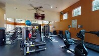 This fitness center is designed to accommodate a wide range of workout routines within a bright, energizing environment. The space features soaring ceilings accented by a bold orange and cream color scheme, creating a motivating atmosphere throughout. Large windows positioned at multiple heights along the walls flood the interior with natural light, while ceiling fans provide added air circulation during workouts.

The strength-training area is comprehensively outfitted with True and TAG brand cable machines, a multi-station functional trainer, leg press and extension machines, adjustable weight benches, a full dumbbell rack, and a medicine ball storage rack stocked with an assortment of weighted balls and stability balls. A wall-mounted flat-screen television is positioned for easy viewing from the strength equipment area.

The dedicated cardio section includes True brand recumbent and upright stationary bikes, a StairMaster elliptical, treadmills, and rowing machines, all arranged to take advantage of the natural light and outdoor views through the floor-to-ceiling glass doors that open directly to an outdoor pool area. The durable rubber flooring throughout provides cushioning and traction. Large mirrors along one wall further enhance the sense of openness within the space.