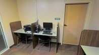 This single-floor space functions as a compact shared computing area designed for practical, task-focused use. The workstation features a continuous desk surface equipped with at least one desktop computer and monitor, a multifunction printer, and a keyboard and mouse setup, providing essential tools for basic computing needs. Dark wood-toned privacy partitions divide the desk space, offering users a degree of separation while working. Two black chairs with metal frames provide seating at the workstation. The flooring consists of patterned carpet tiles in muted tones, contributing a utilitarian aesthetic consistent with a shared-use environment. The walls are finished in a neutral beige tone, keeping the space visually straightforward and functional. A wood-finish door to the right is labeled "Janitor," indicating adjacency to building service facilities. Overhead, visible conduit runs along the wall, reflecting a no-frills, practical design approach. The space is modest in scale, catering to users who need quick access to computing and printing resources without requiring a dedicated private office.