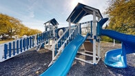 Located at 8115 Worthington Galena Rd in Westerville, Ohio, this outdoor playground offers a comprehensive recreational space designed for active play. The centerpiece of the area is a large, multi-level play structure finished in a striking blue and white color scheme. The structure features covered tower platforms connected by walkways, railings, and bridges, providing children with an engaging environment to explore. Multiple blue slides of varying styles are integrated into the design, including a wide straight slide, a dual side-by-side slide, and a spiraling corkscrew slide that adds an exciting element to the play experience. A textured climbing wall with blue handholds offers an additional physical challenge for older children. Separate from the main structure, a swing set with blue bucket seats provides options for younger visitors. The entire play area is surfaced with gravel for safety, bordered by neat stone edging that defines the playground boundaries. The surrounding landscape includes mature deciduous trees and open grassy areas, adding a natural, park-like setting to the space. Seating benches and a lamp post are positioned nearby, making the area functional and inviting for supervising adults. Parking is accessible adjacent to the playground.