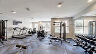 Located at 8011 Augusta Pines Dr in Spring, Texas, this fitness center offers a well-appointed, single-floor exercise facility designed to support a full range of workout routines. The space is finished with a distinctive herringbone-patterned carpet tile flooring complemented by interlocking foam mat sections that define dedicated workout zones, providing both style and functionality underfoot.

Natural light streams in through multiple windows fitted with blinds, creating a bright and energizing atmosphere throughout. Recessed ceiling lighting further ensures consistent illumination across the entire floor area. A wall-mounted flat-screen television is positioned for visibility from the cardio section, enhancing the workout experience.

The cardio zone is equipped with multiple treadmills and elliptical trainers, as well as stationary and cycling bikes. The strength training area features a multi-station cable and pulley machine, a dedicated weight bench, and a full dumbbell rack offering a wide range of free weights. A large floor-to-ceiling mirror spans one wall, visually expanding the space and allowing users to monitor their form during exercises.

The facility is accessed through glass-paneled doors, and the neutral gray walls, clean white trim, and modern equipment combine to create a professional, motivating environment. The overall design is practical and inviting, catering to a variety of fitness needs.