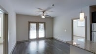 This single-story space is thoughtfully designed with a clean, modern aesthetic that balances comfort and functionality. The main living area is immediately inviting, showcasing rich, dark hardwood flooring that runs throughout and provides a striking contrast against the neutral, light-toned walls. A ceiling fan with an integrated light fixture is centrally positioned overhead, contributing to both air circulation and ambient lighting.

A standout feature of the living area is the pair of French doors, framed with warm wood trim and fitted with horizontal blinds. These doors flood the room with natural light while offering a seamless visual connection to the outdoors. The open layout allows for flexible furniture arrangements and creates a sense of spaciousness.

Adjacent to the living space, a partial view of a kitchen counter reveals granite or stone-look countertops, hinting at quality finishes carried throughout the home. The transition between spaces is smooth and open, reinforcing the flow of the floor plan.

Overall, the space presents a well-maintained interior with consistent finishes, practical design choices, and an atmosphere that feels both bright and grounded — well-suited for everyday living.