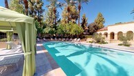 This inviting outdoor pool area exudes a resort-like atmosphere, combining elegant Spanish-inspired architecture with lush, tropical landscaping. The centerpiece is a generously sized swimming pool with crystal-clear turquoise water, surrounded by a spacious tiled deck that offers ample room for relaxation and movement. Towering date palms and mature shade trees frame the entire space, creating a sense of seclusion and natural beauty beneath open blue skies.

Lounge chairs are arranged along the pool deck, providing plentiful sunbathing and relaxation options. A standout feature is the shaded cabana area, where sage-green canopied structures with draped curtains offer a private retreat from the sun, each furnished with comfortable lounge chairs and side tables for an elevated poolside experience.

Access to the pool is secured through a decorative wrought-iron gate framed by a graceful stucco arch, reinforcing the Spanish Colonial architectural character seen throughout the surrounding buildings. Manicured landscaping, including ornamental shrubs and potted plants, adds lush greenery around the perimeter. The combination of warm stucco exteriors, arched architectural details, and abundant palm trees creates a cohesive, resort-caliber ambiance ideal for outdoor leisure and recreation.