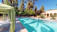 This inviting outdoor pool area exudes a resort-like atmosphere, combining elegant Spanish-inspired architecture with lush, tropical landscaping. The centerpiece is a generously sized swimming pool with crystal-clear turquoise water, surrounded by a spacious tiled deck that offers ample room for relaxation and movement. Towering date palms and mature shade trees frame the entire space, creating a sense of seclusion and natural beauty beneath open blue skies.

Lounge chairs are arranged along the pool deck, providing plentiful sunbathing and relaxation options. A standout feature is the shaded cabana area, where sage-green canopied structures with draped curtains offer a private retreat from the sun, each furnished with comfortable lounge chairs and side tables for an elevated poolside experience.

Access to the pool is secured through a decorative wrought-iron gate framed by a graceful stucco arch, reinforcing the Spanish Colonial architectural character seen throughout the surrounding buildings. Manicured landscaping, including ornamental shrubs and potted plants, adds lush greenery around the perimeter. The combination of warm stucco exteriors, arched architectural details, and abundant palm trees creates a cohesive, resort-caliber ambiance ideal for outdoor leisure and recreation.