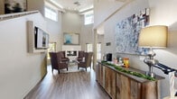 Located at 1660 Drew Circle in Davis, California, this two-floor amenity space offers a well-appointed layout designed for both communal gathering and focused work. The ground floor makes an immediate impression with a soaring double-height ceiling in the main lounge, where large clerestory windows flood the area with natural light. A sleek fireplace anchors the seating area, complemented by wood-look flooring and curated artwork. Adjacent to the lounge, a rustic wood-paneled counter serves as a refreshment station, while a separate dining area features a bistro-style table with views to the outdoors. The kitchen is fully equipped with stainless steel appliances, dark cabinetry, and a generous white-countertop island with bar seating — ideal for social gatherings. A hallway connects the main areas and provides access to the staircase leading to the upper floor. The second floor houses additional living rooms with wall-mounted televisions, plush carpeting, and ample natural light from multiple windows, providing flexible spaces for lounging or media use. A private office rounds out the upper level, outfitted with a full workstation, filing storage, and a printer — a practical retreat for focused tasks. Throughout, the design maintains a cohesive, modern aesthetic with neutral tones and thoughtful finishes.