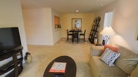 This single-floor space offers a comfortable and inviting layout designed for everyday living. The open-concept design seamlessly connects the living and dining areas, creating a sense of spaciousness and easy flow throughout. The living area is anchored by plush, neutral-toned carpeting that extends throughout the main space, complemented by soft, warm wall colors that lend a welcoming ambiance. A rounded coffee table serves as a central gathering point, accompanied by a cushioned sofa with accent pillows, perfect for relaxed lounging. Recessed lighting illuminates the space from above, while natural light filters in through a nearby window, brightening the interior. The dining area features a dark-toned bistro-style table with matching chairs, positioned beneath a framed decorative art piece that adds a touch of personality to the warm yellow accent wall. A tall ladder-style shelf beside the dining area provides both functional storage and a stylish display surface for personal items and décor. A pass-through window or built-in ledge along one wall hints at a nearby kitchen, adding to the practical flow of the layout. Overall, the space strikes a balance between casual comfort and thoughtful design.