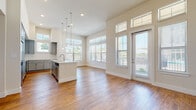 This single-floor unit showcases a thoughtfully designed open-concept layout that seamlessly connects the kitchen, dining, and living areas. The kitchen serves as the centerpiece, featuring a large white-quartz island with an undermount sink, sleek gray shaker cabinetry, stainless steel appliances including a French door refrigerator, and elegant pendant lighting suspended from soaring ceilings. Rich hardwood-style flooring runs throughout the main living areas, complementing the neutral, light-toned walls.

The living space is bathed in natural light, courtesy of oversized windows fitted with horizontal blinds and a glass exterior door with transom windows above, offering outdoor views and easy access. A ceiling fan provides additional comfort in this generously proportioned area.

The bedrooms are carpeted for warmth and comfort, each featuring large windows that invite ample daylight. Walk-in closets with wire shelving systems provide practical storage solutions. The bathroom is finished with gray cabinetry, a white vanity countertop, hardwood-style flooring, and a glass-enclosed shower, maintaining the clean, contemporary aesthetic found throughout the unit. Overall, the space exudes a bright, modern ambiance with consistent, high-quality finishes.