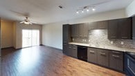 This single-floor unit showcases a thoughtfully designed layout with a cohesive modern aesthetic carried throughout each space. The open kitchen and living area form the heart of the unit, where rich hardwood-style flooring extends across the main living space. The kitchen is outfitted with dark wood cabinetry, granite-style countertops, a textured tile backsplash, track lighting, and black appliances including a refrigerator, range, microwave, and dishwasher. A sliding glass door in the living area leads to a private balcony, allowing natural light to fill the space.

The bedrooms feature plush carpet flooring and ceiling fans, creating a comfortable, quiet atmosphere. Stylish gray panel doors add a contemporary touch, and the primary bedroom connects to a spacious walk-in closet with built-in shelving and hanging rods on multiple walls. The bathroom is finished with light tile flooring, a dark vanity with a granite-style countertop, a large mirror with overhead lighting, a wall-mounted cabinet, and a shower-tub combination. Dark baseboards throughout the unit tie together the overall modern design.