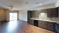 This single-floor unit showcases a thoughtfully designed layout with a cohesive modern aesthetic carried throughout each space. The open kitchen and living area form the heart of the unit, where rich hardwood-style flooring extends across the main living space. The kitchen is outfitted with dark wood cabinetry, granite-style countertops, a textured tile backsplash, track lighting, and black appliances including a refrigerator, range, microwave, and dishwasher. A sliding glass door in the living area leads to a private balcony, allowing natural light to fill the space.

The bedrooms feature plush carpet flooring and ceiling fans, creating a comfortable, quiet atmosphere. Stylish gray panel doors add a contemporary touch, and the primary bedroom connects to a spacious walk-in closet with built-in shelving and hanging rods on multiple walls. The bathroom is finished with light tile flooring, a dark vanity with a granite-style countertop, a large mirror with overhead lighting, a wall-mounted cabinet, and a shower-tub combination. Dark baseboards throughout the unit tie together the overall modern design.