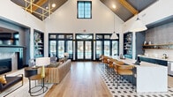 Located at 1528 Balch Rd in Madison, Alabama, this single-story community clubhouse offers a generously sized, open-plan interior designed with a sophisticated modern-farmhouse aesthetic. The space is anchored by dramatic vaulted ceilings with exposed wood beams and dark-painted shiplap, creating an impressive sense of height and volume throughout.

The main gathering area features warm hardwood-style flooring that transitions to a bold geometric-patterned tile beneath the central bar island. A large white quartz island with leather-upholstered bar stools serves as a social hub, flanked by a full wet bar with dark cabinetry, open wood shelving, and a textured tile backsplash. Oversized pendant lights illuminate the space from above.

A comfortable lounge area is anchored by a teal-trimmed fireplace with an elegant tile surround, a wall-mounted television, and built-in shelving with bold patterned wallpaper accents. Plush seating and stylish accent tables complete this relaxed setting.

The leasing office area is enclosed with sleek glass-and-steel partitions, maintaining visual openness while providing a dedicated workspace. Dark-framed French doors and floor-to-ceiling windows flood the interior with natural light and frame views of the outdoor pool and surrounding grounds, seamlessly connecting the interior to the exterior environment.