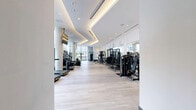This professionally designed fitness facility spans a single floor and comprises two dedicated exercise rooms, together offering a comprehensive and well-appointed workout environment. The main exercise room is expansive and flooded with natural light through floor-to-ceiling windows that frame lush tropical foliage, creating an energizing atmosphere. Light hardwood-style flooring runs throughout, complementing the crisp white walls and elegant coffered ceilings with warm recessed and cove lighting. The space is equipped with a full lineup of premium cardio machines, including treadmills, ellipticals, and steppers, arranged along the windowed perimeter. A dedicated strength training zone features cable machines, weight benches, plate-loaded equipment, and a full dumbbell rack, with large decorative mirrors lining the walls to enhance depth and visibility. Wall-mounted flat-screen televisions provide entertainment throughout. The second exercise room offers a more intimate setting, fitted with a multi-functional RAX-style training station, a heavy bag, a speed bag, resistance training accessories, a stability ball, and kettlebells — ideal for functional and high-intensity training. Light-toned flooring and pendant lighting give this room a clean, focused ambiance. The facility entrance features sleek double glass doors and a controlled access entry, contributing to the overall polished, modern aesthetic of the space.