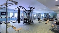 This fitness center is designed to support a full range of workout routines, offering an impressive selection of professional-grade equipment within a single, open floor layout. At the heart of the space stands a Hoist multi-functional cable and strength training machine, complemented by a heavy punching bag for combat-style training. A dedicated free weights area features a bench and a full rack of dumbbells, while cardio machines — including treadmills and an elliptical — line the perimeter for endurance training. The room is finished with light-toned, resilient flooring that provides both durability and comfort underfoot. Soft blue walls lend a calm, motivating atmosphere, while large windows along multiple walls invite generous natural light into the space. Full-length mirrors are mounted along one wall, enhancing the sense of openness and allowing users to monitor their form. Ceiling fans are strategically placed overhead to maintain air circulation throughout a workout session. Recessed lighting supplements the natural light, ensuring the space remains bright and functional at any time of day. The overall ambiance is clean, well-organized, and purpose-built for effective fitness training.