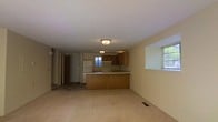 This two-floor residential space offers a functional and straightforward layout designed for comfortable everyday living. The main level showcases an open-concept design where the living area flows seamlessly into the kitchen, creating a spacious and connected environment. The kitchen is outfitted with warm wood-toned cabinetry, a white refrigerator, a range with an overhead exhaust, and a sink set along a peninsula-style counter. The peninsula serves as a natural divider between the kitchen and the living area, adding both utility and definition to the space. Soft carpeting covers the living area floor, contributing to a cozy atmosphere, while flush-mounted ceiling light fixtures provide consistent overhead illumination throughout. Multiple windows allow natural light to filter into the space, brightening the neutral-toned walls and creating an airy feel. A hallway visible from the main living area leads to additional rooms and access points, suggesting a well-organized floor plan. The second floor adds further living space, expanding the overall capacity of the home. The clean, understated finishes throughout give the space a versatile, move-in-ready character suitable for a variety of living arrangements.