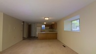 This two-floor residential space offers a functional and straightforward layout designed for comfortable everyday living. The main level showcases an open-concept design where the living area flows seamlessly into the kitchen, creating a spacious and connected environment. The kitchen is outfitted with warm wood-toned cabinetry, a white refrigerator, a range with an overhead exhaust, and a sink set along a peninsula-style counter. The peninsula serves as a natural divider between the kitchen and the living area, adding both utility and definition to the space. Soft carpeting covers the living area floor, contributing to a cozy atmosphere, while flush-mounted ceiling light fixtures provide consistent overhead illumination throughout. Multiple windows allow natural light to filter into the space, brightening the neutral-toned walls and creating an airy feel. A hallway visible from the main living area leads to additional rooms and access points, suggesting a well-organized floor plan. The second floor adds further living space, expanding the overall capacity of the home. The clean, understated finishes throughout give the space a versatile, move-in-ready character suitable for a variety of living arrangements.