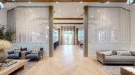 Located at 7100 Summerside Dr in Whitsett, NC, this thoughtfully designed single-floor community space blends modern farmhouse aesthetics with upscale finishes throughout. The open-concept layout is anchored by striking light-toned herringbone hardwood floors that flow seamlessly from one zone to the next, creating a cohesive and inviting atmosphere.

Soaring ceilings with exposed wood beams add architectural warmth and volume, while sheer floor-to-ceiling drapery panels flank structural columns at the entrance, framing the space with an elegant, airy quality. Natural light pours in through oversized windows and glass French doors, enhancing the bright and welcoming ambiance.

The living area features multiple distinct seating arrangements, including a cozy lounge anchored by a floor-to-ceiling whitewashed brick fireplace topped with a mounted television and flanked by framed artwork. An arched ceiling detail overhead adds dramatic architectural interest. A separate sitting area showcases a sculptural 3D wall treatment, paired with eclectic furnishings and a statement chandelier.

The kitchen bar area is outfitted with a marble-topped island, black Windsor barstools, lantern pendant lighting, and exposed brick accent walls. A full-height black refrigerator and glass-paneled French doors leading to an outdoor area complete the functional yet stylish kitchen zone. The space as a whole conveys a refined yet relaxed character, ideal for community gathering and social interaction.