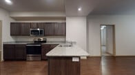 Located at 1534 N Olympic Club Ave in Fayetteville, Arkansas, this single-story home offers a thoughtfully designed layout with consistent finishes throughout. The kitchen serves as a central highlight, featuring dark espresso-toned cabinetry, granite countertops, and a full suite of stainless steel appliances including a range, microwave, and refrigerator. A peninsula with an integrated sink and additional counter space adds both functionality and a natural gathering point that opens toward the rest of the living area. Hardwood-style flooring runs throughout the main living and kitchen areas, lending warmth and a cohesive aesthetic. The bedrooms are carpeted for comfort, each equipped with ceiling fans and closet space; the primary bedroom notably includes a walk-in closet alongside an additional secondary closet. Windows with blinds allow natural light into the bedrooms while maintaining privacy. The bathrooms reflect the same design language found in the kitchen, with dark wood vanities, light stone-topped counters, and hardwood-style flooring. The primary bath features a dual-sink vanity. The floor plan, as seen in the dollhouse view, flows efficiently between living, sleeping, and utility spaces, making this a practical and well-finished single-level home.