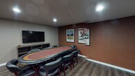 Located at 250 Pacific Ave in Long Beach, California, this single-floor entertainment space is thoughtfully designed for social gatherings and leisure. The room is anchored by a full-size oval poker table surrounded by cushioned leather chairs, making it well-suited for card games and game nights. A large flat-screen television is mounted on the wall adjacent to a media console, providing additional entertainment options for guests.

The lounge area features a coordinated arrangement of black leather armchairs, a sofa, and a round coffee table, creating a comfortable space for conversation and relaxation. Recessed lighting throughout the space provides consistent, ambient illumination, while the dark hardwood-style flooring and textured wall coverings contribute to a moody, upscale aesthetic. Vintage-style whiskey advertisement posters accent the warm, terra cotta-toned feature wall, adding a distinctive character to the room.

A kitchenette is integrated into the space, equipped with dark wood cabinetry, a sink with stone-look countertops, and a compact refrigerator, offering convenient amenities for hosting. A wall-mounted split air conditioning unit ensures climate control. Glass-paneled entry doors connect the space to an adjacent corridor, providing easy accessibility. Overall, the space delivers a refined, club-like atmosphere suited for entertaining.