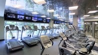 This fitness center offers a comprehensive and well-appointed workout environment designed to support an active lifestyle. The space is dominated by an impressive lineup of cardio equipment, including treadmills, elliptical trainers, and additional aerobic machines arranged in orderly rows to maximize both capacity and usability.

Large floor-to-ceiling windows line the perimeter of the room, flooding the space with abundant natural light and offering lush, tree-filled views that create a refreshing and motivating atmosphere during workouts. A striking mirrored ceiling runs the length of the room, amplifying the sense of openness and adding a sleek, modern aesthetic to the environment.

A continuous row of flat-screen televisions is mounted along the upper walls, providing entertainment and news content across multiple channels — ensuring members can stay informed and engaged throughout their sessions. Recessed panel lighting further enhances the bright and energetic ambiance of the space.

The flooring is finished in durable, dark rubberized material, standard for high-traffic fitness environments and designed to support both comfort and safety. Overall, this fitness center combines modern design, ample natural light, and a full suite of cardio equipment to deliver a premium exercise experience.