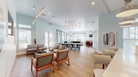 Located at Mason Pond Place in Ladson, South Carolina, this single-floor community clubhouse offers a generously sized, open-plan gathering space designed for both relaxation and social engagement. The interior is defined by soaring ceilings with white shiplap detailing, light wood-look flooring throughout, and a soft, airy color palette of pale blue-gray and crisp white that lends the space a coastal, welcoming character.

The layout flows seamlessly across several distinct functional zones. A comfortable lounge area features modern upholstered seating arrangements, wall-mounted televisions, and curated artwork, while expansive windows and multiple sets of glass French doors flood the space with natural light and offer views of the surrounding community. Elegant pendant chandeliers and contemporary light fixtures serve as architectural focal points overhead.

A centrally located dining area with a large communal table and modern black chairs sits beneath a striking cluster of globe pendant lights. Adjacent to this is a game zone equipped with a foosball table and a full shuffleboard table, ideal for recreational use.

The fully appointed kitchen features floor-to-ceiling gray shaker cabinetry with glass-front upper panels, quartz countertops, stainless steel appliances including a built-in oven and full-size refrigerator, and a large center island with bar seating — making this space well-suited for community events and gatherings.