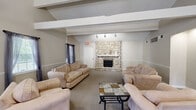 Located at 4583 Whisper Lake Dr in Black Jack, Missouri, this single-floor community clubhouse offers a well-appointed gathering space designed for both relaxation and recreation. The layout encompasses three distinct areas that flow cohesively throughout the facility. The main lounge is anchored by a striking floor-to-ceiling natural stone fireplace with a white mantle, serving as the room's focal point. Exposed ceiling beams add architectural character, while neutral-toned carpeting and wainscoting along the walls contribute to a warm, welcoming ambiance. Comfortable seating arrangements invite residents to gather and unwind. A dedicated game room features soaring vaulted ceilings with exposed beams and houses a full-size pool table with pendant lighting overhead, complemented by additional seating for spectators. A glass-paneled door provides access to an adjacent office or utility area. The third area functions as a dining and transitional space, furnished with a glass-top dining table and wrought iron chairs beneath vaulted ceilings with track lighting. A secondary lounge nook nearby features a built-in display alcove with recessed lighting and a decorative console table. Throughout, the space maintains a consistent design palette of neutral walls, carpeted floors, and classic wainscoting, creating a cohesive and functional community environment.