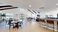 Located at 111 Sunset Cir in Camden, DE, this single-floor community space offers an expansive, open-concept layout that seamlessly blends a fully equipped kitchen with a generously sized living area. The architectural design is highlighted by dramatic vaulted ceilings adorned with rustic exposed wood beams, creating a warm yet modern farmhouse aesthetic throughout.

The living area is thoughtfully arranged into multiple functional zones, including a lounge with a large wall-mounted television, plush leather seating, and accent chairs arranged around a patterned area rug. A shuffleboard table and pool table add a recreational dimension to the space. A dedicated dining area features a round wooden table with cross-back chairs, positioned near glass French doors that allow natural light to flood the room and provide views of the surrounding community.

The kitchen is designed for large-scale use, featuring an expansive U-shaped layout with warm natural wood cabinetry, bold dark countertops, a stainless steel sink, dishwasher, and a beverage refrigerator. A bar-height counter with leather stools connects the kitchen to the living space, encouraging a social atmosphere.

Light oak hardwood flooring runs throughout, complementing the crisp white walls and recessed lighting. Large windows on multiple sides of the space invite ample daylight and exterior views, reinforcing an open, airy ambiance ideal for community gatherings.