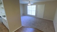 This single-floor residential space offers a clean and functional layout designed for comfortable everyday living. The main living area is a highlight of the space, featuring plush wall-to-wall carpeting that transitions seamlessly into hardwood-style flooring near the entryway, creating a clear and practical delineation between zones. A ceiling fan with an integrated light fixture is centrally mounted, providing both air circulation and ambient lighting throughout the room. A large window along the far wall allows generous amounts of natural light to fill the space, lending an open and airy feel to the interior. The entry door, positioned adjacent to the window, features a classic six-panel design with a standard deadbolt, offering a welcoming yet secure point of entry. The walls are finished in a neutral, warm beige tone, complemented by clean white baseboards and trim throughout. A floor-level vent is visible along the baseboard, indicating a standard forced-air climate control system. The textured ceiling adds a subtle traditional character to the overall aesthetic. The space presents a straightforward, move-in-ready environment with practical finishes suited for a wide range of living arrangements.