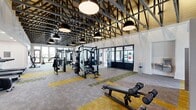 Located at 170 Graphics Dr in Madison, Alabama, this expansive fitness center offers a generously sized, single-floor layout designed to accommodate a full range of workout needs. The architectural design is immediately striking, with soaring exposed steel truss ceilings accented by warm wood paneling and oversized industrial pendant lighting, creating a dynamic, open-air atmosphere throughout the main exercise floor.

The primary workout area is outfitted with an extensive array of strength training machines, including multi-station cable systems, weight benches, and a full dumbbell rack neatly organized behind a slatted room divider. Cardio equipment — including treadmills, air bikes, a curved HIIT treadmill, and rowing machines — lines the space, with large floor-to-ceiling windows and glass doors along the perimeter flooding the area with natural light and providing views of the surrounding outdoor landscape.

Flooring throughout the main area features a patterned carpet tile in shades of gold, green, and gray, adding visual interest and defining workout zones. A dedicated studio room offers hardwood flooring, full-wall mirrors, Peloton-style bikes, a rowing machine, a wall-mounted TV, and expansive dark-framed windows, making it ideal for cycling and functional training. A water fountain and ample open floor space complete this well-appointed facility.
