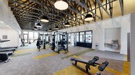 Located at 170 Graphics Dr in Madison, Alabama, this expansive fitness center offers a generously sized, single-floor layout designed to accommodate a full range of workout needs. The architectural design is immediately striking, with soaring exposed steel truss ceilings accented by warm wood paneling and oversized industrial pendant lighting, creating a dynamic, open-air atmosphere throughout the main exercise floor.

The primary workout area is outfitted with an extensive array of strength training machines, including multi-station cable systems, weight benches, and a full dumbbell rack neatly organized behind a slatted room divider. Cardio equipment — including treadmills, air bikes, a curved HIIT treadmill, and rowing machines — lines the space, with large floor-to-ceiling windows and glass doors along the perimeter flooding the area with natural light and providing views of the surrounding outdoor landscape.

Flooring throughout the main area features a patterned carpet tile in shades of gold, green, and gray, adding visual interest and defining workout zones. A dedicated studio room offers hardwood flooring, full-wall mirrors, Peloton-style bikes, a rowing machine, a wall-mounted TV, and expansive dark-framed windows, making it ideal for cycling and functional training. A water fountain and ample open floor space complete this well-appointed facility.