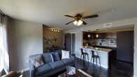 This single-floor space showcases a thoughtfully designed open-concept layout that seamlessly integrates the living, dining, and kitchen areas into one cohesive environment. The living area is anchored by a plush gray sectional sofa accented with decorative pillows, complemented by a ceiling fan with integrated lighting that provides both comfort and ambiance. Dark hardwood flooring runs throughout, adding a sleek, modern aesthetic that ties the spaces together.

A cozy dining nook is positioned adjacent to the living area, featuring a compact round table suited for intimate gatherings. The kitchen is a standout feature, boasting dark cabinetry, stainless steel appliances, and a granite-topped breakfast bar with industrial-style bar seating — ideal for casual dining or entertaining. Pendant lighting above the bar area adds a warm, inviting glow, while recessed lighting enhances the kitchen's functionality.

The neutral gray wall tones and accent wall with decorative artwork contribute to a contemporary, urban atmosphere. A hallway visible from the main living space suggests additional private rooms beyond, completing this well-appointed and efficiently designed single-level residence.