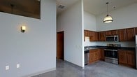 This single-story space makes a strong architectural impression through its dramatically tall ceilings, which lend an open, airy quality to the interior. The polished concrete floors run continuously throughout, providing a sleek, industrial aesthetic that complements the clean white walls and crisp lines of the space.

The kitchen is positioned along one wall and is defined by warm medium-toned wood cabinetry that extends both above and below the dark countertops, offering generous storage. A stainless steel sink is centrally placed beneath upper cabinets, and stainless steel appliances, including a dishwasher and range, integrate seamlessly into the layout. A pendant light fixture above the kitchen area adds a functional and decorative touch.

A structural column and partial wall divide the kitchen from the adjacent open living area, creating a subtle sense of zone separation while preserving the spacious feel. A doorway visible from the main area leads to additional rooms beyond. High windows near the ceiling allow natural light to filter in from above, enhancing the vertical drama of the space. Overall, the design conveys a modern, utilitarian character with clean, unfussy finishes throughout.